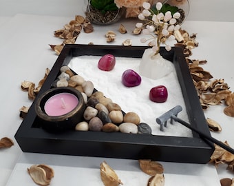 May include: A miniature zen garden in a black wooden tray. It features white sand, polished stones, and three red-purple stones. A small candle in a bronze-colored holder and a miniature rake are also included. A small tree with pink stones is in the background.