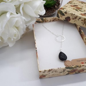 May include: A silver necklace with a teardrop-shaped black pendant is displayed inside a floral patterned gift box. The necklace has a circular design and a delicate chain. White roses are in the background.
