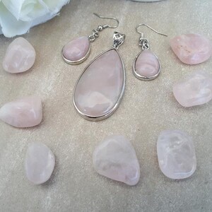 Puede incluir: Un conjunto de joyas y piedras de cuarzo rosa. El conjunto incluye un colgante en forma de l&aacute;grima, pendientes a juego y varias piedras de cuarzo rosa pulidas. Las joyas tienen engastes de metal plateado. Las piedras son de color rosa.
