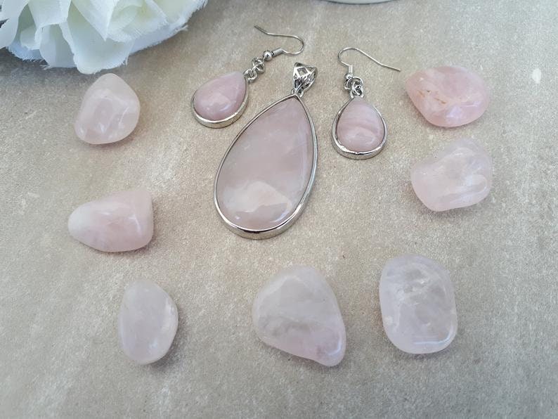 Puede incluir: Un conjunto de joyas y piedras de cuarzo rosa. El conjunto incluye un colgante en forma de l&aacute;grima, pendientes a juego y varias piedras de cuarzo rosa pulidas. Las joyas tienen engastes de metal plateado. Las piedras son de color rosa.