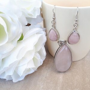 Puede incluir: Un conjunto de joyas con un colgante y pendientes a juego. El colgante y los pendientes tienen forma de l&aacute;grima y son de color rosa p&aacute;lido, engastados en metal plateado. Los pendientes tienen cierre de gancho. Flores blancas en el fondo.