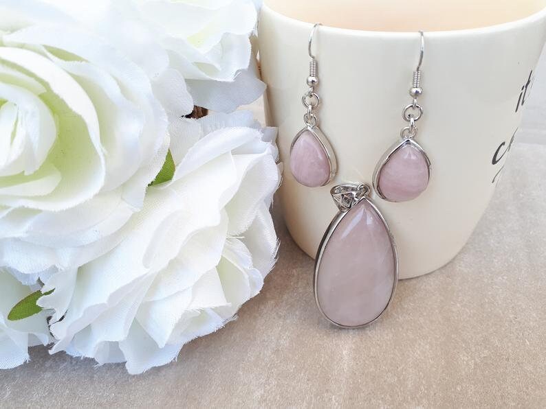 Puede incluir: Un conjunto de joyas con un colgante y pendientes a juego. El colgante y los pendientes tienen forma de l&aacute;grima y son de color rosa p&aacute;lido, engastados en metal plateado. Los pendientes tienen cierre de gancho. Flores blancas en el fondo.