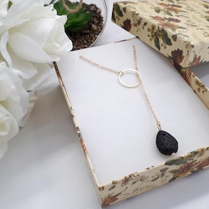 Puede incluir: Un collar dorado con un colgante circular y un colgante de piedra de lava negra en forma de lágrima. El collar se muestra dentro de una caja de regalo con estampado floral. Rosas blancas y vegetación en el fondo.