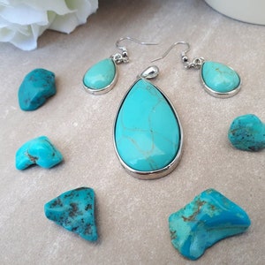 Puede incluir: Un conjunto de joyas de turquesa con un colgante en forma de l&aacute;grima y pendientes a juego, todo engarzado en plata. Varias piedras de turquesa en bruto est&aacute;n dispersas alrededor de las joyas. La turquesa tiene un color azul-verdoso vibrante con patrones marrones.