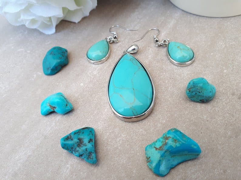 Puede incluir: Un conjunto de joyas de turquesa con un colgante en forma de l&aacute;grima y pendientes a juego, todo engarzado en plata. Varias piedras de turquesa en bruto est&aacute;n dispersas alrededor de las joyas. La turquesa tiene un color azul-verdoso vibrante con patrones marrones.