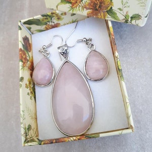 Puede incluir: Un conjunto de joyas con un colgante en forma de l&aacute;grima y pendientes a juego, todo en un tono rosa suave. El conjunto est&aacute; engastado en metal plateado y se presenta en una caja de regalo con estampado floral. Los pendientes y el colgante tienen un acabado liso y pulido.