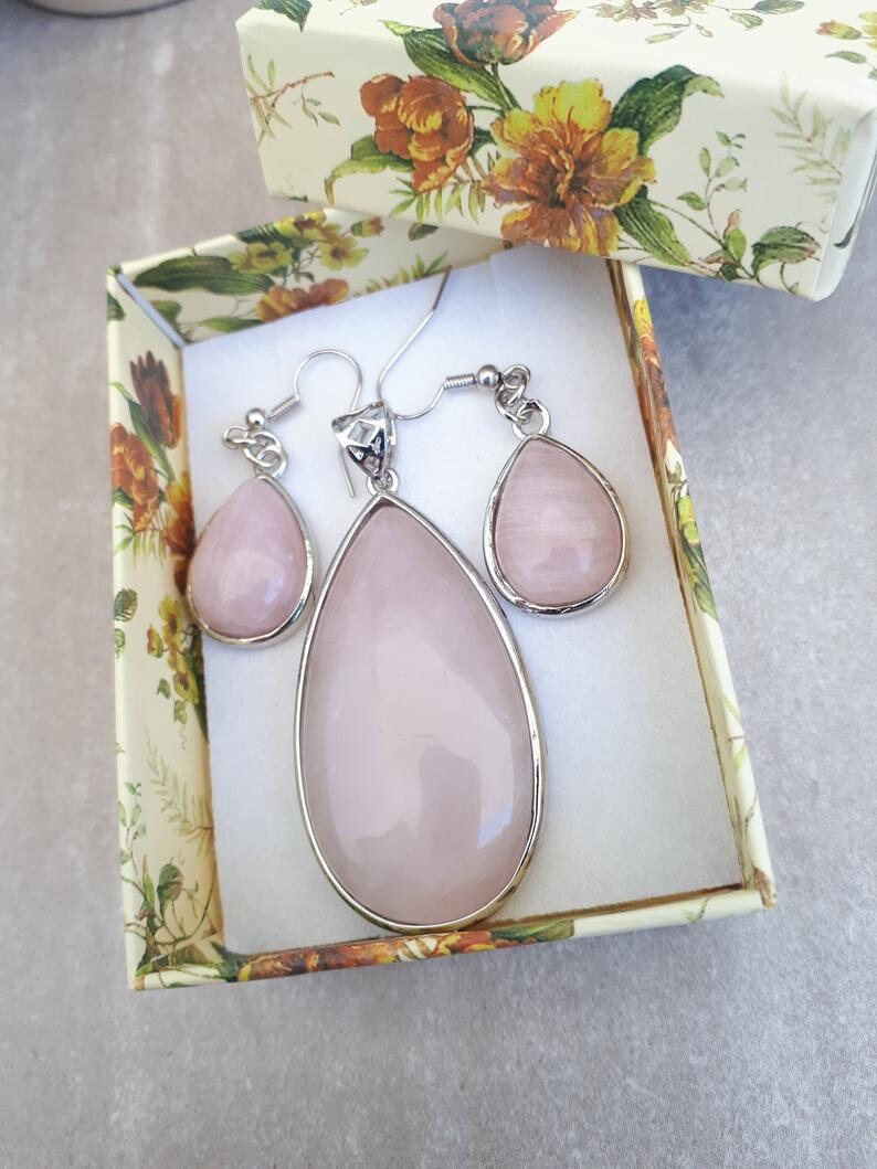 Puede incluir: Un conjunto de joyas con un colgante en forma de l&aacute;grima y pendientes a juego, todo en un tono rosa suave. El conjunto est&aacute; engastado en metal plateado y se presenta en una caja de regalo con estampado floral. Los pendientes y el colgante tienen un acabado liso y pulido.