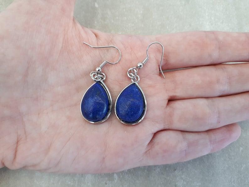 Pu&ograve; includere: Un paio di orecchini a goccia con pietre blu scuro incastonate in metallo color argento. Gli orecchini sono mostrati su una mano, evidenziando le loro dimensioni e il design. I ganci sono color argento.
