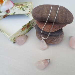 Könnte beinhalten: Eine silberne Halskette mit einem tropfenförmigen Rosenquarz-Anhänger liegt auf einem Steinhaufen. Mehrere lose Rosenquarzsteine und eine florale Geschenkbox sind ebenfalls vorhanden.