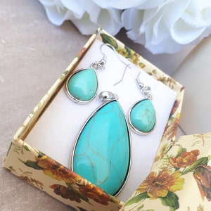 Puede incluir: Un conjunto de joyas turquesas, que incluye un colgante y pendientes a juego, presentado en una caja de regalo con estampado floral. El colgante y los pendientes tienen un borde de metal plateado. Las piedras turquesas tienen un aspecto veteado.