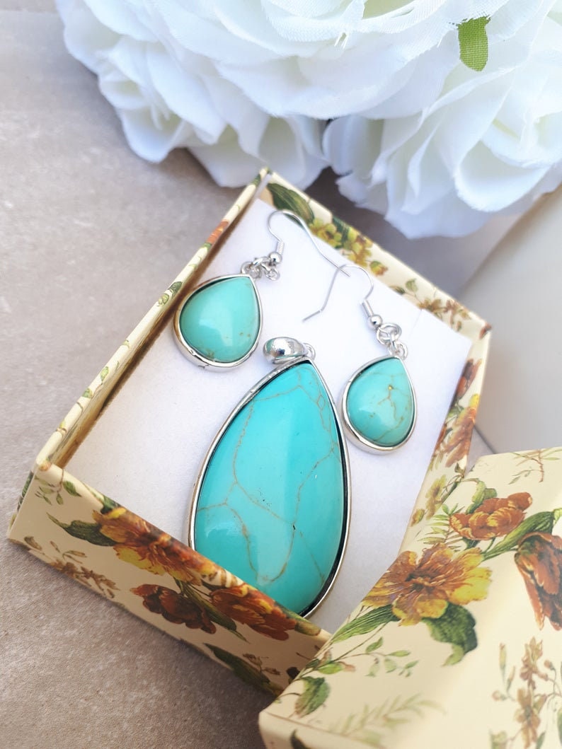 Puede incluir: Un conjunto de joyas turquesas, que incluye un colgante y pendientes a juego, presentado en una caja de regalo con estampado floral. El colgante y los pendientes tienen un borde de metal plateado. Las piedras turquesas tienen un aspecto veteado.