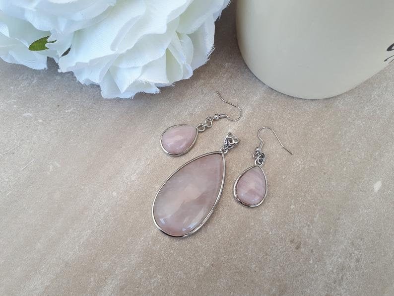 Puede incluir: Un conjunto de joyas de cuarzo rosa con un colgante y pendientes a juego. El colgante y los pendientes tienen forma de l&aacute;grima y est&aacute;n engarzados en metal plateado. El cuarzo rosa tiene un tono rosa suave. El conjunto est&aacute; sobre una superficie neutra.