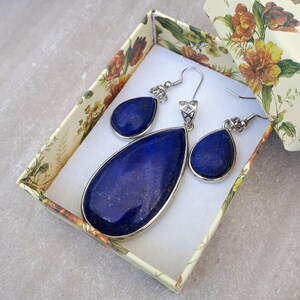 Pu&ograve; includere: Un set di gioielli con un ciondolo e orecchini abbinati, presentato in una scatola decorativa floreale. Il ciondolo e gli orecchini presentano grandi pietre blu scuro a forma di goccia con montature in metallo color argento. La scatola ha uno sfondo crema con motivi floreali.