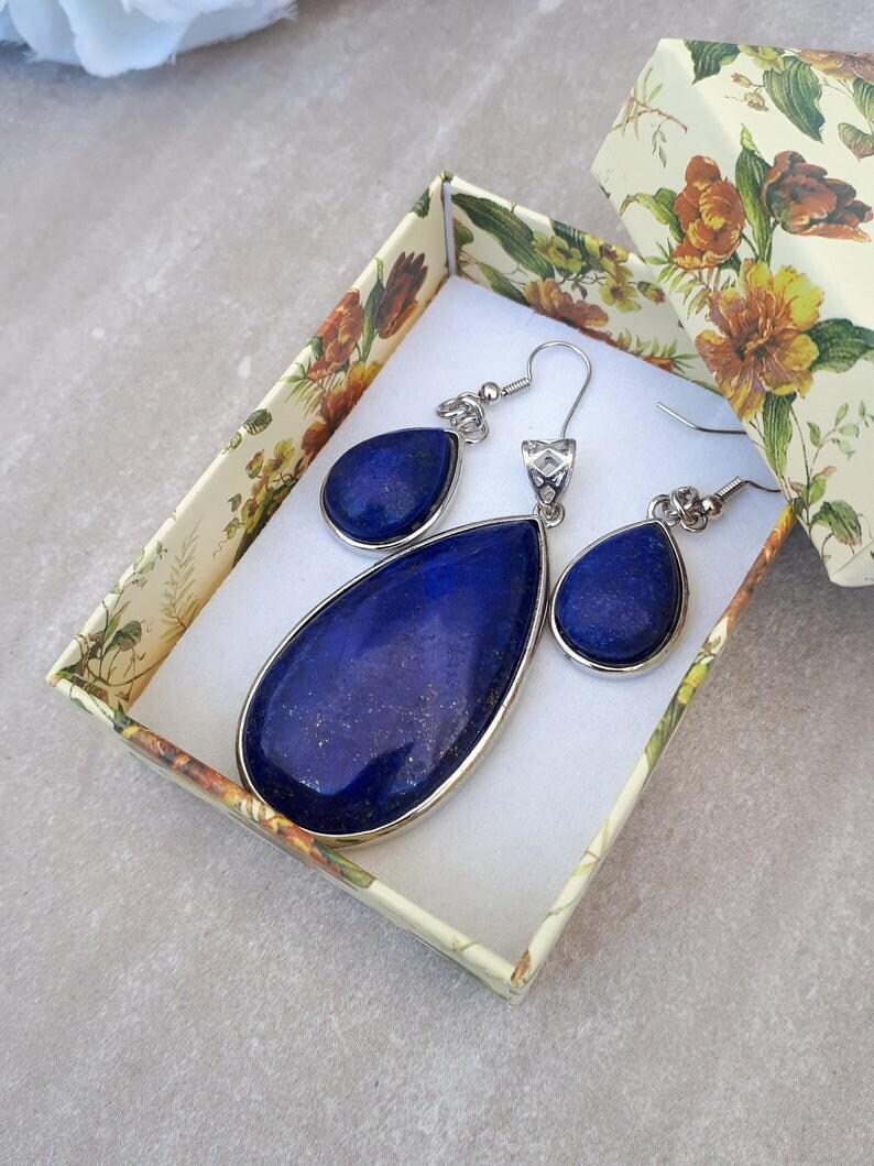 Pu&ograve; includere: Un set di gioielli con un ciondolo e orecchini abbinati, presentato in una scatola decorativa floreale. Il ciondolo e gli orecchini presentano grandi pietre blu scuro a forma di goccia con montature in metallo color argento. La scatola ha uno sfondo crema con motivi floreali.