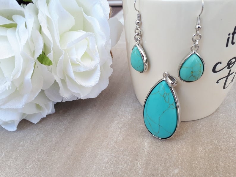 Puede incluir: Un conjunto de joyas turquesas con un colgante y pendientes a juego. Las piedras en forma de l&aacute;grima est&aacute;n engastadas en metal plateado. Los pendientes tienen cierre de gancho. Flores blancas en el fondo.