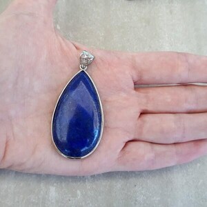 Pu&ograve; includere: Un ciondolo a forma di goccia con una pietra blu scuro incastonata in una cornice di metallo color argento. Il ciondolo ha una piccola cauzione decorativa nella parte superiore. La pietra &egrave; di un blu intenso e il ciondolo &egrave; tenuto in una mano.