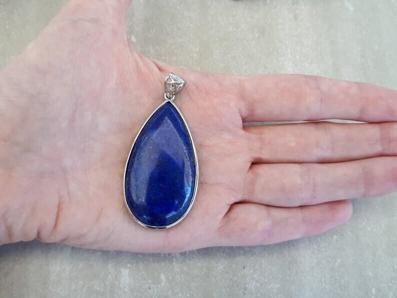 Pu&ograve; includere: Un ciondolo a forma di goccia con una pietra blu scuro incastonata in una cornice di metallo color argento. Il ciondolo ha una piccola cauzione decorativa nella parte superiore. La pietra &egrave; di un blu intenso e il ciondolo &egrave; tenuto in una mano.