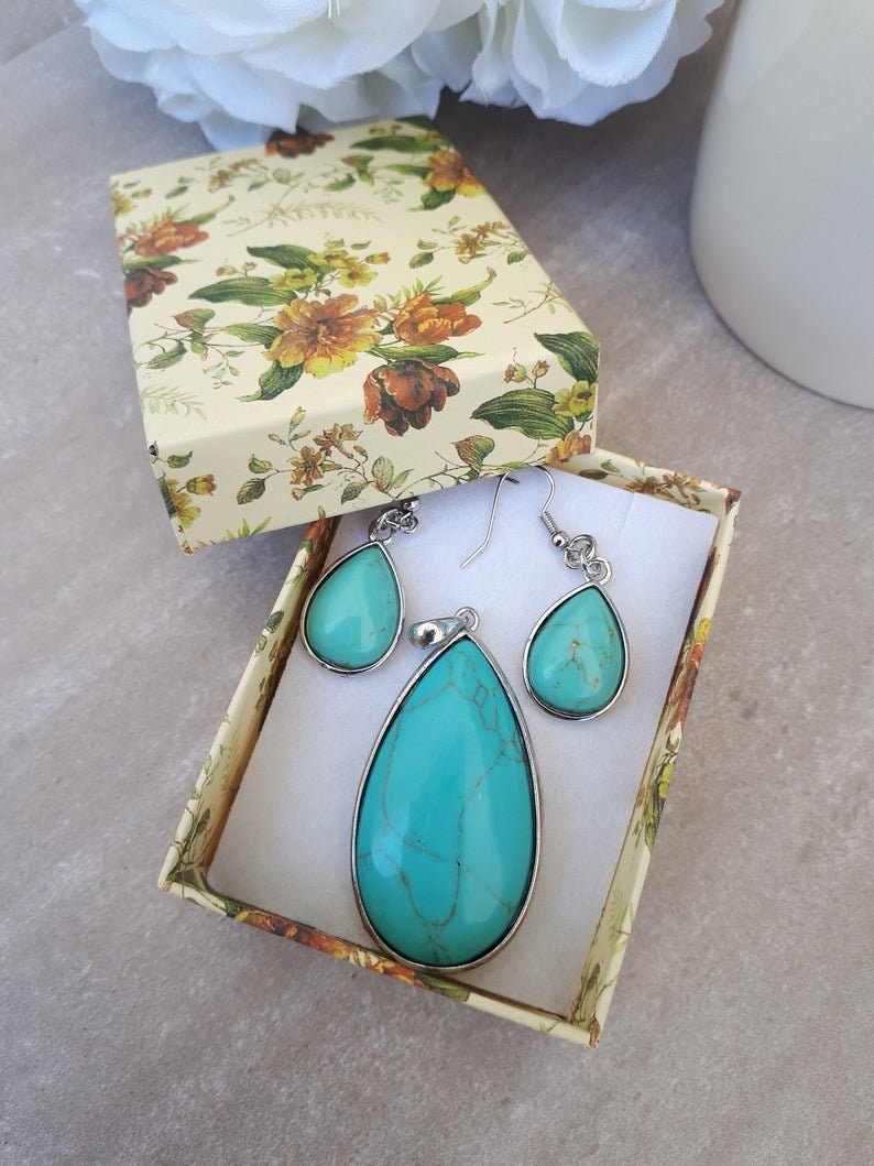 Puede incluir: Un conjunto de joyas turquesas, que incluye un colgante y pendientes, presentado en una caja de regalo con estampado floral. Las piedras en forma de l&aacute;grima est&aacute;n engastadas en metal plateado. La caja est&aacute; abierta, revelando las joyas.