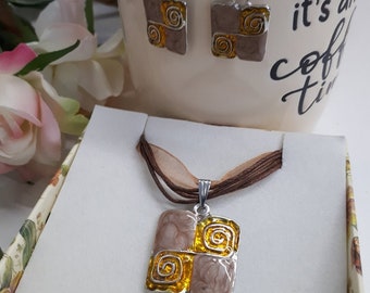 May include: A jewelry set featuring a pendant and matching earrings. The pendant and earrings have a square design with a two-tone color scheme of brown and gold, with a spiral design. The set is presented in a decorative box.