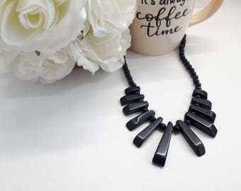 Puede incluir: Un collar negro con colgantes geométricos. El collar se muestra con una taza de café que dice "It's always coffee time" y rosas blancas.
