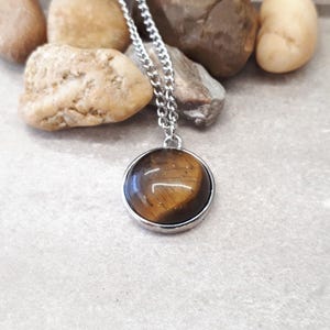 May include: A silver-toned necklace with a round pendant featuring a polished tiger's eye stone. The pendant is set in a silver bezel and hangs from a delicate chain. The necklace is displayed on a light gray surface with small rocks.