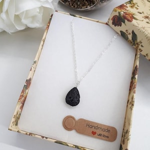 May include: A silver chain necklace with a black teardrop-shaped pendant, presented in a floral patterned gift box. A small brown tag with the words "Handmade with love" is also in the box. The box is open, revealing a white interior.
