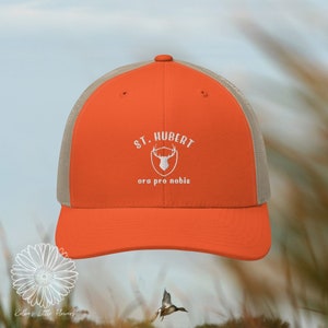 May include: Orange and beige trucker hat with a white embroidered logo that says "St. Hubert oro pro nobis" and features a deer head inside a shield.