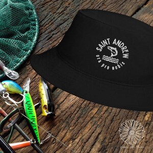 May include: Black bucket hat with a white embroidered logo that says "Saint Andrew Ora Pro Nobis" and a jumping fish inside a circle.