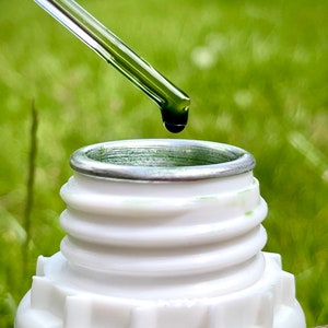 May include: A clear glass dropper with a green liquid is being used to add a drop of liquid to a white bottle with a silver lid. The bottle is sitting on a green grassy surface.
