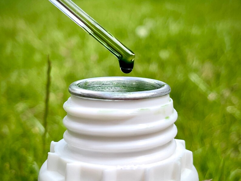 May include: A clear glass dropper with a green liquid is being used to add a drop of liquid to a white bottle with a silver lid. The bottle is sitting on a green grassy surface.