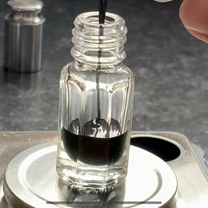 May include: A clear glass bottle with a black liquid inside, sitting on a silver scale. A thin black stick is inserted into the bottle.