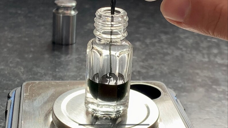 May include: A clear glass bottle with a black liquid inside, sitting on a silver scale. A thin black stick is inserted into the bottle.