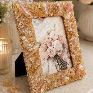 May include: A rectangular picture frame covered in small, golden-yellow crystal chips and larger, elongated, translucent crystal points. The frame holds a photograph of a bouquet of pale pink and white peonies with green stems and lace fabric.