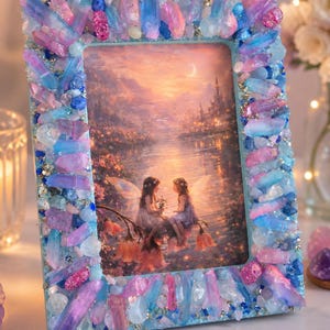 May include: A rectangular picture frame decorated with an array of blue and pink crystals and small stones. The frame holds an image of two winged figures sitting on flowers near a lake with a castle in the background under a crescent moon.