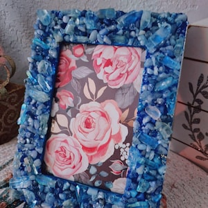 May include: A rectangular photo frame with a blue crystal-encrusted border. The frame holds a floral print featuring pink roses on a dark background. The frame is a decorative item.