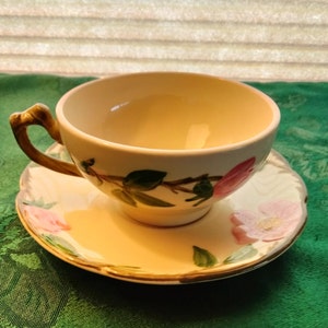 May include: A vintage teacup and saucer set with a floral design featuring pink roses and green leaves. The cup has a brown handle and the saucer has a gold rim.