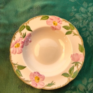 May include: A white ceramic soup bowl with a floral design. The bowl features pink roses with green leaves and stems. The bowl is on a green tablecloth.