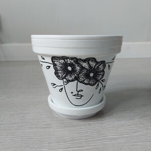 May include: White ceramic planter featuring a black and white illustration of a face with flowers covering the eyes. The flowers are stylised and have a simple design.