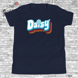 May include: A navy blue t-shirt with a retro-style graphic of the word "Daisy" in white with a blue daisy flower in the center. The word is outlined in rainbow colors.