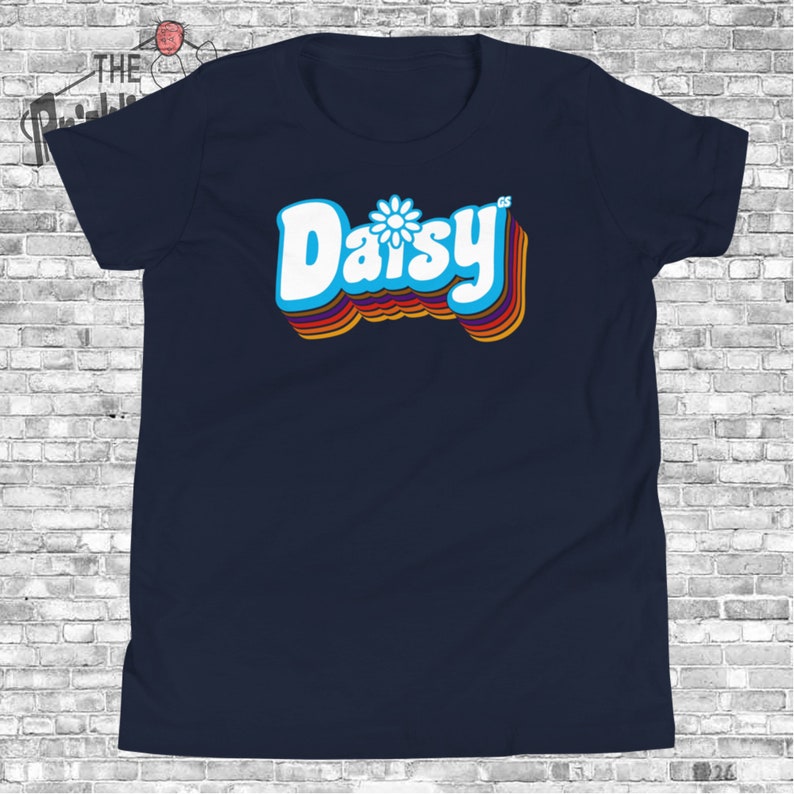 May include: A navy blue t-shirt with a retro-style graphic of the word "Daisy" in white with a blue daisy flower in the center. The word is outlined in rainbow colors.