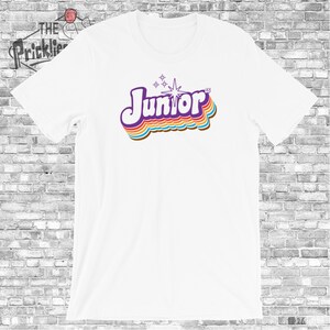May include: White t-shirt with a retro-style graphic of the word "Junior" in purple with rainbow-colored stripes and a white star.