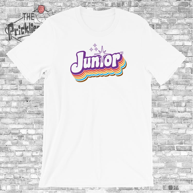 May include: White t-shirt with a retro-style graphic of the word "Junior" in purple with rainbow-colored stripes and a white star.