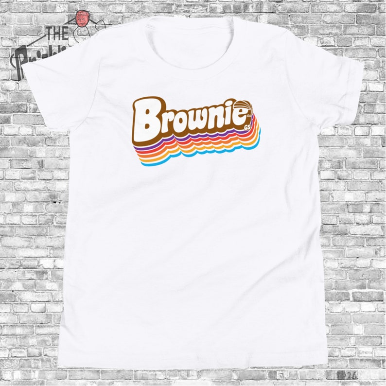 May include: White t-shirt with a retro-style graphic of the word "Brownie" in brown letters with rainbow stripes behind it.