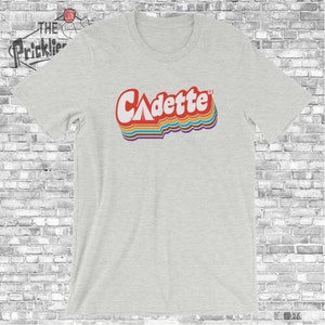 K&ouml;nnte beinhalten: Ein hellgraues T-Shirt mit einer Retro-Grafik des Wortes "Cadette" mit einem Zelt-Symbol im "A".