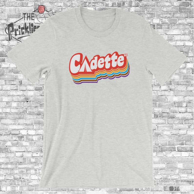 K&ouml;nnte beinhalten: Ein hellgraues T-Shirt mit einer Retro-Grafik des Wortes "Cadette" mit einem Zelt-Symbol im "A".