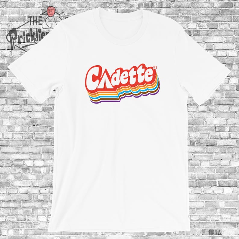 K&ouml;nnte beinhalten: Wei&szlig;es T-Shirt mit einer Retro-Grafik des Wortes "Cadette" in Regenbogenfarben. Das Wort "Cadette" enth&auml;lt ein stilisiertes Zelt-Symbol im "C".