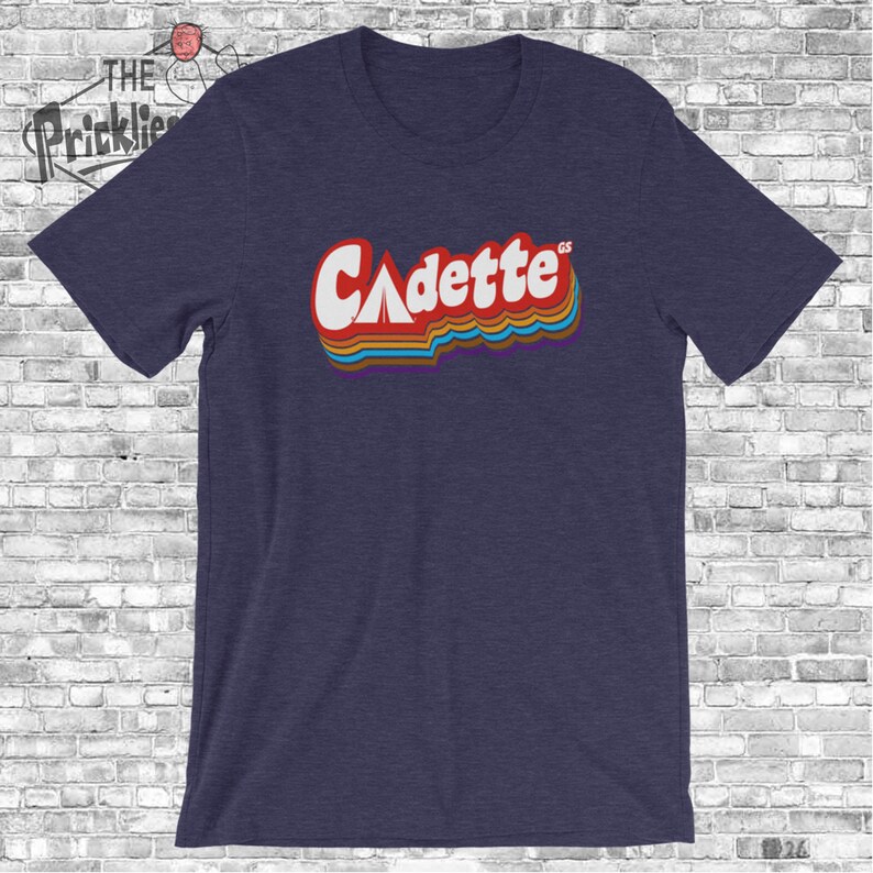 K&ouml;nnte beinhalten: Ein marineblaues T-Shirt mit einer Retro-Grafik des Wortes "Cadette" mit einem Zelt-Symbol und den Buchstaben "GS" in kleinerer Schrift.