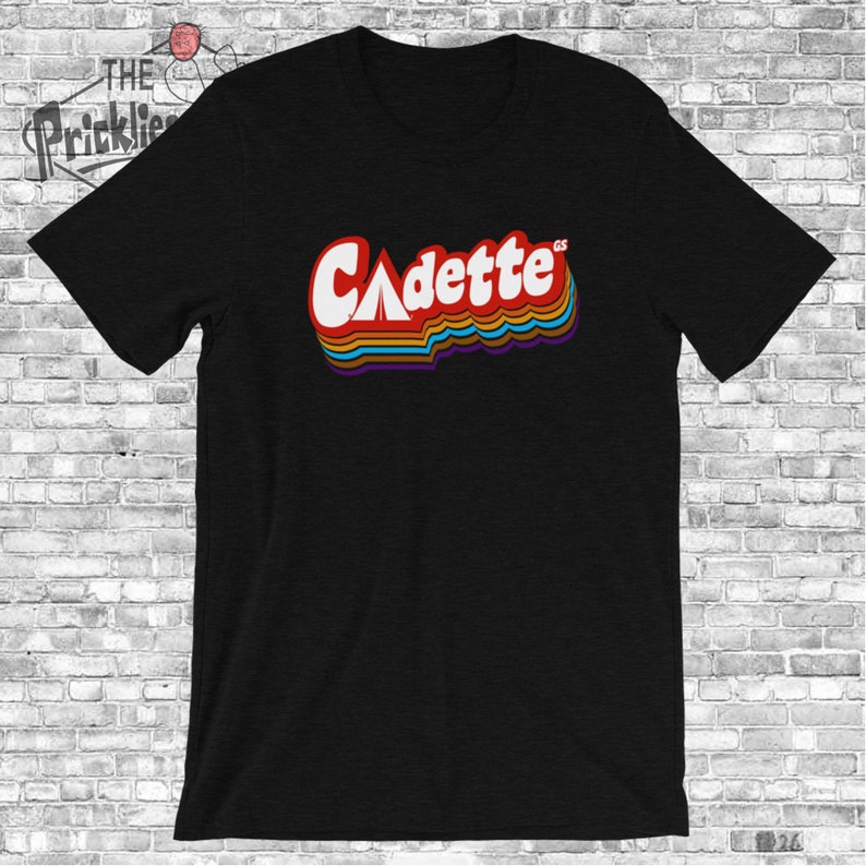 K&ouml;nnte beinhalten: Schwarzes T-Shirt mit einer Retro-Grafik des Wortes "Cadette" in Regenbogenfarben. Das Wort "GS" steht in kleineren Buchstaben &uuml;ber dem Wort "Cadette".