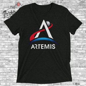 Puede incluir: Una camiseta negra con el logotipo de Artemis. El logotipo es una letra A blanca con una línea roja que la atraviesa y un círculo azul alrededor. La palabra "ARTEMIS" está escrita debajo del logotipo.