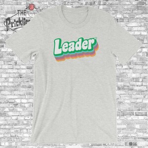 May include: A heather grey t-shirt with a retro-style graphic of the word "Leader" in rainbow colors.